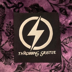 May include: A square black fabric patch featuring a white graphic of a lightning bolt enclosed within a circle. Below the circular emblem, the text "THROBBING GRISTLE" is printed in a stylised white font. The patch has a visible woven texture.