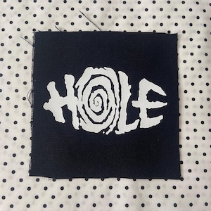May include: A square black fabric patch featuring the word HOLE printed in white, with the letter O stylised as a spiral. The patch has raw, slightly frayed edges on the left side and a woven texture visible across the dark surface.