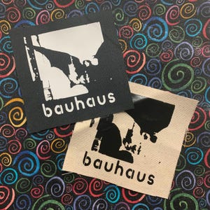 May include: Two square fabric patches featuring the word "bauhaus". One is black with a white graphic, the other is beige with a black graphic. The background is a colourful swirl pattern. The patches are for clothing.