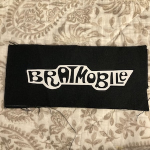 May include: A black rectangular patch with the white text "BRATMOBILE" in a stylized font. The patch has a rough, unfinished edge and is set against a patterned fabric background.