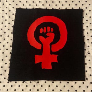以下が含まれることがあります： 黒い正方形の布製パッチで、鮮やかな赤色のグラフィックプリントが施されています。デザインは円の中に拳を突き上げた形をしており、下に向かって十字の形に伸びています。パッチの側面には、ほつれた縁が見えています。