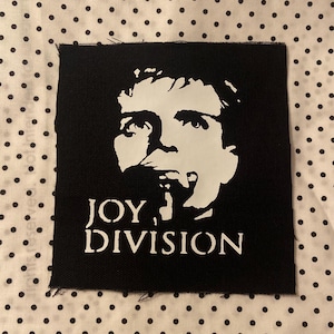 May include: A square black fabric patch featuring a white screen-printed graphic of a person's face and the text JOY DIVISION printed in white capital letters below the portrait. The patch has raw, slightly frayed edges on all four sides.