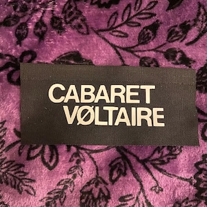 May include: A rectangular black fabric patch featuring the white text CABARET VØLTAIRE in a bold, sans-serif font. The patch has a woven texture and is positioned against a purple background with a dark, intricate floral pattern.