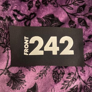 Puede incluir: Un parche rectangular de tela negra con el texto blanco FRONT 242 impreso en una fuente negrita sin remates. El parche está colocado sobre un fondo de tela de felpa morada que muestra un patrón botánico negro repetitivo de hojas y flores.