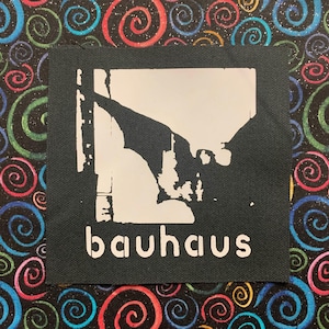 Può includere: Una toppa quadrata nera con un motivo Bauhaus bianco e la parola "bauhaus" in bianco. La toppa è posta su uno sfondo colorato di motivi a spirale.