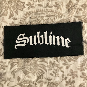 Op de afbeelding: Zwart rechthoekig embleem met het woord "Sublime" in witte gotische letters. De patch is gemaakt van stof en heeft een rafelige rand met zichtbare stiksels. De achtergrond is een stof met bloemenpatroon.
