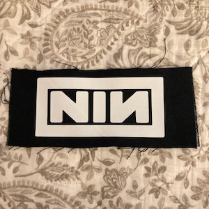Può includere: Toppa rettangolare nera con le lettere bianche "NIN" all'interno di un rettangolo bianco. La toppa è in tessuto e presenta fili sciolti sui bordi. Lo sfondo è un tessuto fantasia.