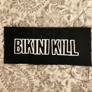 Puede incluir: Un parche rectangular negro con las palabras "BIKINI KILL" en letras mayúsculas blancas. El parche tiene bordes deshilachados y está sobre un fondo de tela estampada.