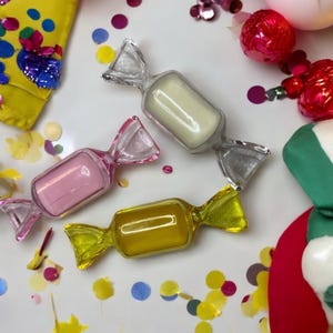 May include: Three clear plastic candy-shaped containers with different coloured fillings. The containers are on a white surface with colourful confetti scattered around them.