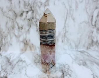 Könnte beinhalten: Ein geschichteter Kristallobelisk mit spitzer Spitze. Der Stein weist Farbbänder auf, darunter Beige, Grau, Schwarz und Lila. Der Kristall steht aufrecht vor einem Marmorhintergrund.