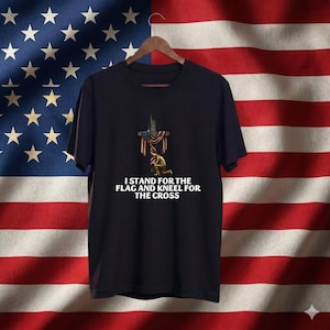 May include: Black t-shirt with the text "I stand for the flag and kneel for the cross." The shirt features a graphic of a cross draped with the American flag and a kneeling soldier. The shirt is hanging on a wooden hanger.