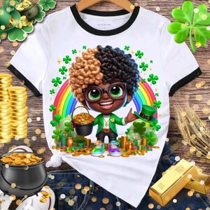 May include: White t-shirt with black trim featuring a cartoon leprechaun with a rainbow, clovers, and a pot of gold. The leprechaun wears glasses, a green jacket, and a green hat. The shirt is on a wooden background.
