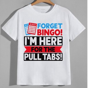 May include: A white short-sleeve t-shirt featuring a graphic print with two bingo cards and the text "FORGET BINGO! I'M HERE FOR THE PULL TABS!" in bold red, black, and blue lettering. The design is centered on the chest of the crew neck garment.
