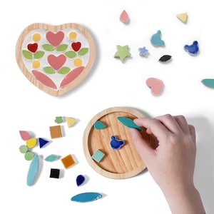 May include: A heart-shaped wooden coaster with a mosaic design of pink and red hearts, green leaves, and yellow circles. Various colourful mosaic tiles, including stars, squares, and teardrops are scattered around. A hand places a blue leaf-shaped tile on a round wooden coaster.