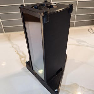 May include: A black, rectangular, 3D-printed object with a triangular base and a clear, rectangular window. The top has four square openings and is secured with screws. The object is on a white surface.