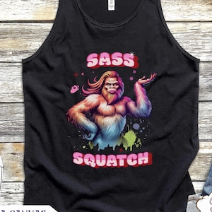 Peut inclure: Débardeur noir avec un graphisme coloré de Sasquatch et le texte "SASS SQUATCH". Le motif représente une créature ressemblant à Bigfoot avec de longs cheveux et une fourrure colorée. Le débardeur est un Bella Canvas Comfy Shirt.