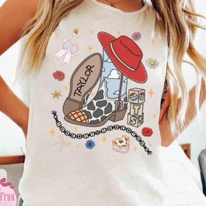 May include: A white sleeveless shirt featuring a graphic print of a red cowboy hat, a cow-print boot with the name Taylor, and stacked blocks with the letters T, S, and the number 13. The design includes a beaded necklace and small floral accents.