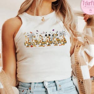 May include: A white tank top featuring a Halloween-themed graphic. The graphic depicts cartoon characters dressed in costumes, holding pumpkins, and surrounded by cobwebs and bats. The top is sleeveless and has a round neckline.