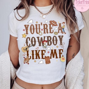 May include: A white short-sleeved t-shirt featuring the brown text "YOU'RE A COWBOY LIKE ME" surrounded by colorful illustrations of a cowboy hat, a guitar, a horse, a boot, stars, clouds, and cartoon characters in a western-themed graphic design.