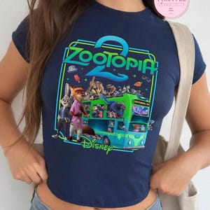 Puede incluir: Top corto azul marino con un gr&aacute;fico colorido de la pel&iacute;cula de Disney "Zootopia 2". El dise&ntilde;o presenta personajes y un autob&uacute;s de dos pisos, con el t&iacute;tulo de la pel&iacute;cula en verde y azul. El logotipo de Disney est&aacute; en la parte inferior.
