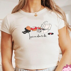 Puede incluir: Camiseta color crema con un gr&aacute;fico de una persona acostada junto a un tocadiscos. La ilustraci&oacute;n incluye el texto "Leave me alone to die". Un disco de vinilo est&aacute; al lado del tocadiscos. La camiseta tiene mangas cortas.