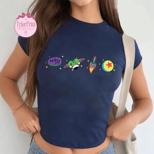 May include: A navy blue cropped t-shirt with a graphic print of characters from the movie Toy Story. The graphic includes a rocket, a ball with a star, a heart-shaped alien, and a purple and green toy with the word "ANDY" on it.