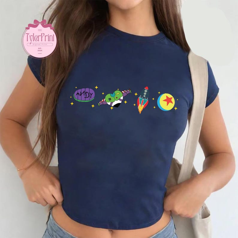May include: A navy blue cropped t-shirt with a graphic print of characters from the movie Toy Story. The graphic includes a rocket, a ball with a star, a heart-shaped alien, and a purple and green toy with the word "ANDY" on it.