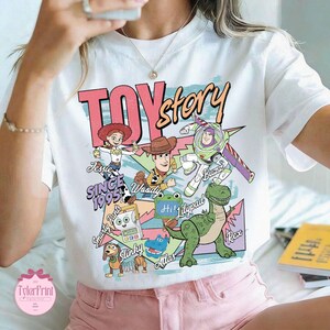 K&ouml;nnte beinhalten: Ein wei&szlig;es T-Shirt mit einem farbenfrohen Grafikdruck der Toy Story-Figuren, darunter Jessie, Woody, Buzz Lightyear, Rex und Slinky. Das Design enth&auml;lt den Text Toy Story, Since 1995 sowie die Namen der Figuren in einem Retro-Stil.