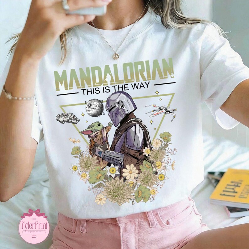 May include: White t-shirt featuring a graphic of The Mandalorian and Grogu, with the text "MANDALORIAN THIS IS THE WAY". The design includes images of the Death Star, a spaceship, and flowers. The shirt is a crew neck.