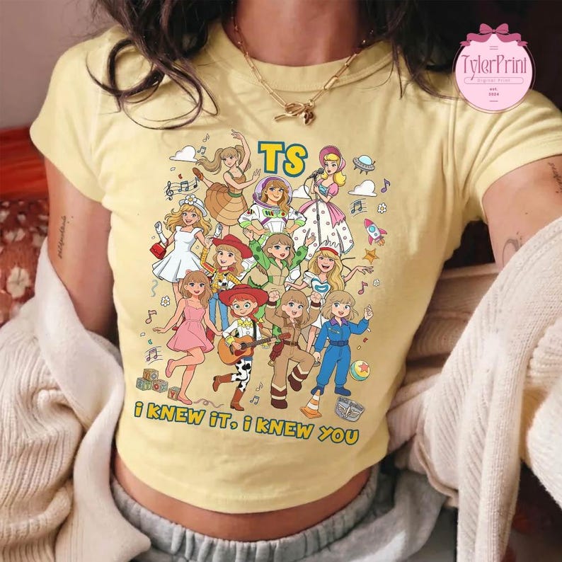 May include: A pale yellow short-sleeve t-shirt featuring a colorful graphic print of various animated characters in a collage style. The text TS is printed at the top, and the text I KNEW IT, I KNEW YOU is printed at the bottom of the graphic.