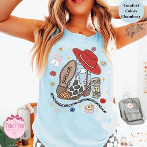 May include: A light blue sleeveless shirt featuring a graphic print of a cow-print cowboy boot, a red cowboy hat, a bow, and stacked blocks with the letters T, S, and S. The name Taylor is written on the boot, and a beaded string design surrounds the graphics.