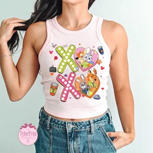 May include: A light pink tank top with a graphic design featuring the characters from Zootopia. The design includes the letters "XO" with heart-shaped illustrations of the characters, coffee cups, and other small icons. The tank top is sleeveless.