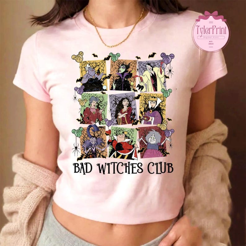 May include: A light pink t-shirt featuring a grid of cartoon villain illustrations. The shirt has the words "BAD WITCHES CLUB" printed in a bold font. The design includes bats, spiders, and Mickey Mouse-shaped balloons.