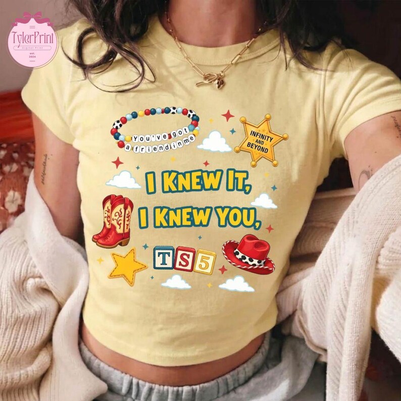 May include: A pale yellow short-sleeve t-shirt featuring a graphic print with a beaded bracelet, a gold sheriff star badge, a red cowboy boot, a red cowboy hat, and alphabet blocks spelling TS5. The text reads I KNEW IT, I KNEW YOU.