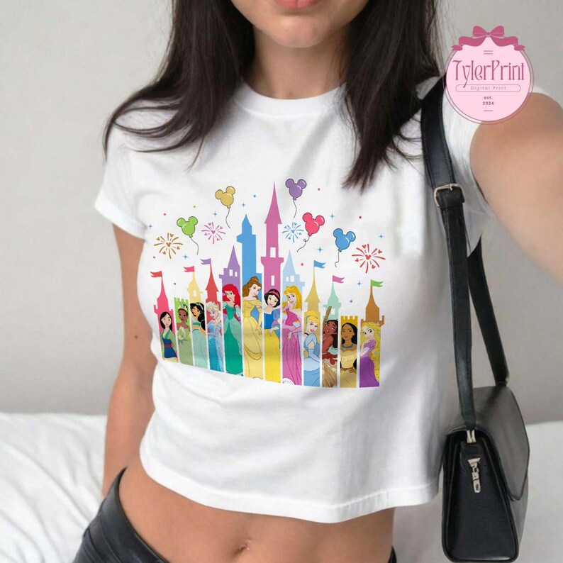 May include: White crop top featuring a colourful graphic of Disney princesses in front of a castle with balloons and fireworks. The text "TylerPrint" is in the top right corner of the image.