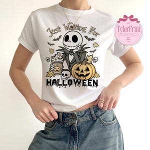Puede incluir: Camiseta blanca con un dise&ntilde;o tem&aacute;tico de Halloween. El dise&ntilde;o incluye un personaje de esqueleto, una calabaza, murci&eacute;lagos y el texto "Just Waiting For Halloween". El personaje sostiene una taza de caf&eacute;. La camiseta es de cuello redondo.