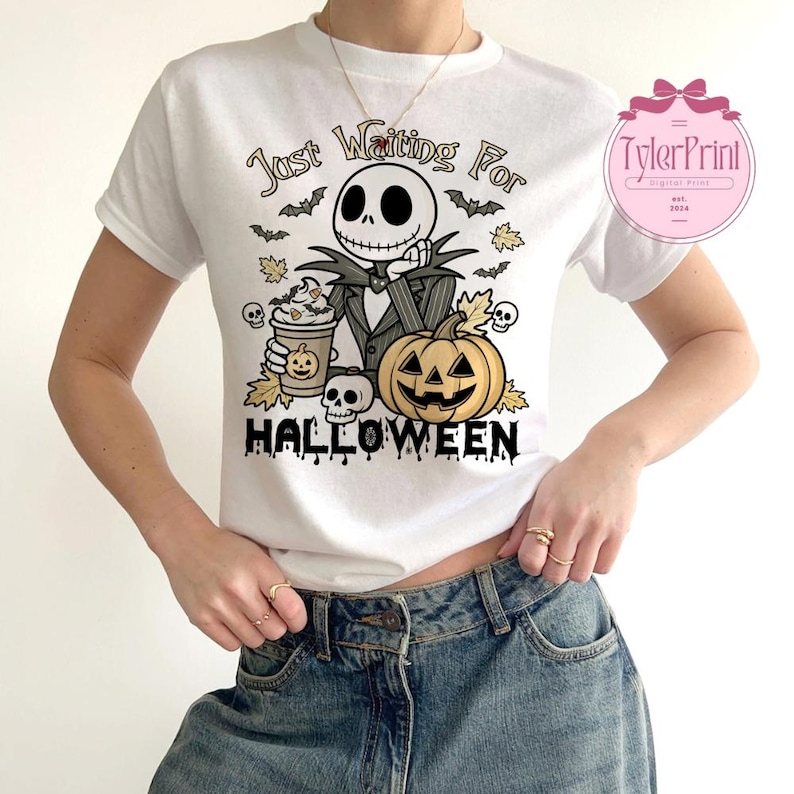 Puede incluir: Camiseta blanca con un dise&ntilde;o tem&aacute;tico de Halloween. El dise&ntilde;o incluye un personaje de esqueleto, una calabaza, murci&eacute;lagos y el texto "Just Waiting For Halloween". El personaje sostiene una taza de caf&eacute;. La camiseta es de cuello redondo.