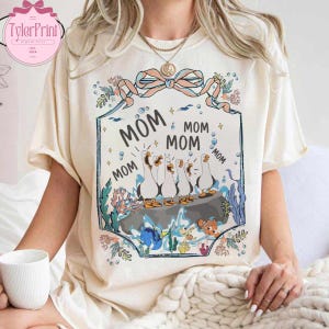 May include: Cream-colored t-shirt featuring a whimsical design with the word "MOM" repeated around a framed illustration. The illustration depicts a group of cartoon geese on a rock, surrounded by aquatic elements. The shirt has short sleeves.