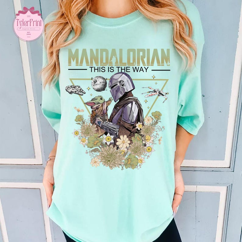 May include: A light blue t-shirt featuring a graphic of The Mandalorian and Grogu surrounded by flowers and space-themed elements. The text "MANDALORIAN - THIS IS THE WAY" is printed across the top. The shirt has a relaxed fit.