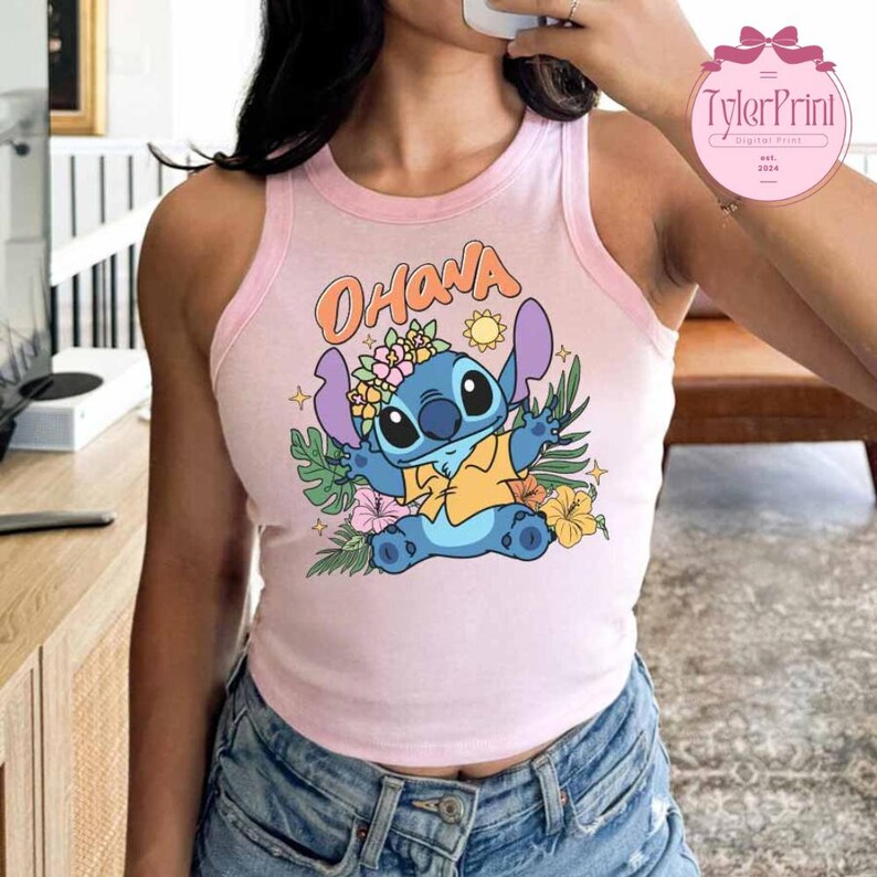 May include: Pink tank top with a light pink trim featuring a cartoon Stitch graphic. The graphic includes the word "Ohana", a sun, flowers, and tropical leaves. The tank top is made of a soft material.