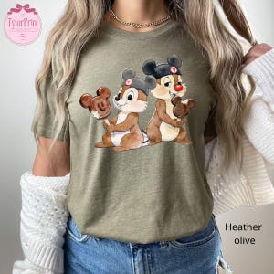 Puede incluir: Camiseta verde oliva jaspeado con una ilustraci&oacute;n de Chip y Chop sosteniendo helados de Mickey Mouse. Los personajes de dibujos animados llevan sombreros. La camiseta tiene el texto "TylerPrint" en la parte superior izquierda. El texto "Heather olive" est&aacute; en la parte inferior derecha.
