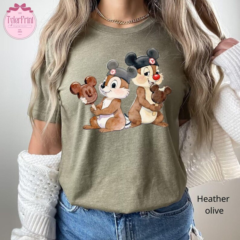 Puede incluir: Camiseta verde oliva jaspeado con una ilustraci&oacute;n de Chip y Chop sosteniendo helados de Mickey Mouse. Los personajes de dibujos animados llevan sombreros. La camiseta tiene el texto "TylerPrint" en la parte superior izquierda. El texto "Heather olive" est&aacute; en la parte inferior derecha.