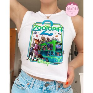Puede incluir: Camiseta sin mangas blanca con un gr&aacute;fico colorido de la pel&iacute;cula animada Zootopia 2. El dise&ntilde;o incluye el t&iacute;tulo de la pel&iacute;cula, personajes y un autob&uacute;s verde y azul. Tambi&eacute;n se ve el logotipo de Disney.