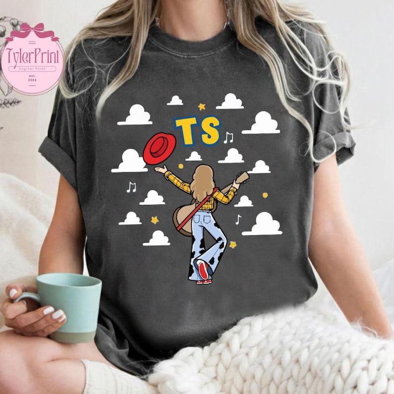 May include: A charcoal gray t-shirt featuring a graphic of a person with long hair wearing a yellow plaid shirt and cow-print bell-bottom pants. The person holds a guitar and a red hat, surrounded by white clouds, yellow stars, and the text TS.