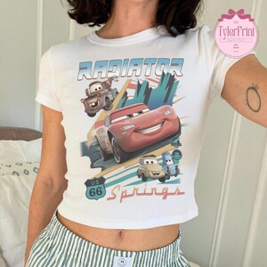 May include: A white short-sleeved t-shirt featuring a graphic print of characters from the movie Cars. The design includes the text Radiator Springs and US 66, with illustrations of Lightning McQueen, Mater, Luigi, and Guido in a desert setting.