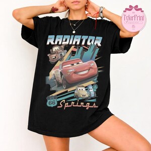 May include: A black short-sleeve t-shirt featuring a graphic print with the text Radiator Springs and US 66. The design includes illustrations of four animated cars in red, brown, and light blue colors set against a stylized background.