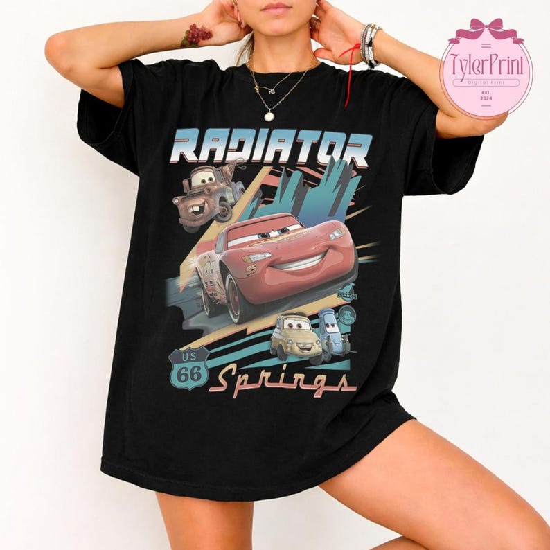 May include: A black short-sleeve t-shirt featuring a graphic print with the text Radiator Springs and US 66. The design includes illustrations of four animated cars in red, brown, and light blue colors set against a stylized background.