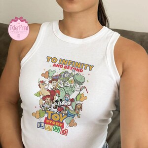 以下が含まれることがあります： 白いタンクトップ。カラフルなトイ・ストーリーのキャラクターのグラフィックが特徴です。「TO INFINITY AND BEYOND」と「TOY STORY LAND」のフレーズが含まれています。タンクトップはノースリーブで、丸いネックラインです。