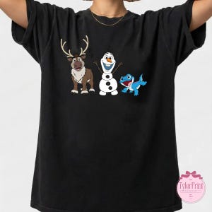 Peut inclure: T-shirt noir avec un dessin anim&eacute; repr&eacute;sentant un renne, un bonhomme de neige et une cr&eacute;ature bleue ressemblant &agrave; un l&eacute;zard. Le renne est brun avec des bois, le bonhomme de neige est blanc et le l&eacute;zard est bleu. Le t-shirt a un col rond.