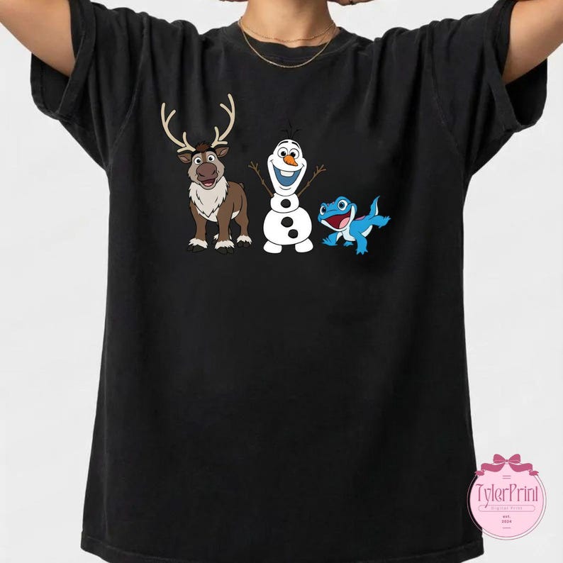 Peut inclure: T-shirt noir avec un dessin anim&eacute; repr&eacute;sentant un renne, un bonhomme de neige et une cr&eacute;ature bleue ressemblant &agrave; un l&eacute;zard. Le renne est brun avec des bois, le bonhomme de neige est blanc et le l&eacute;zard est bleu. Le t-shirt a un col rond.
